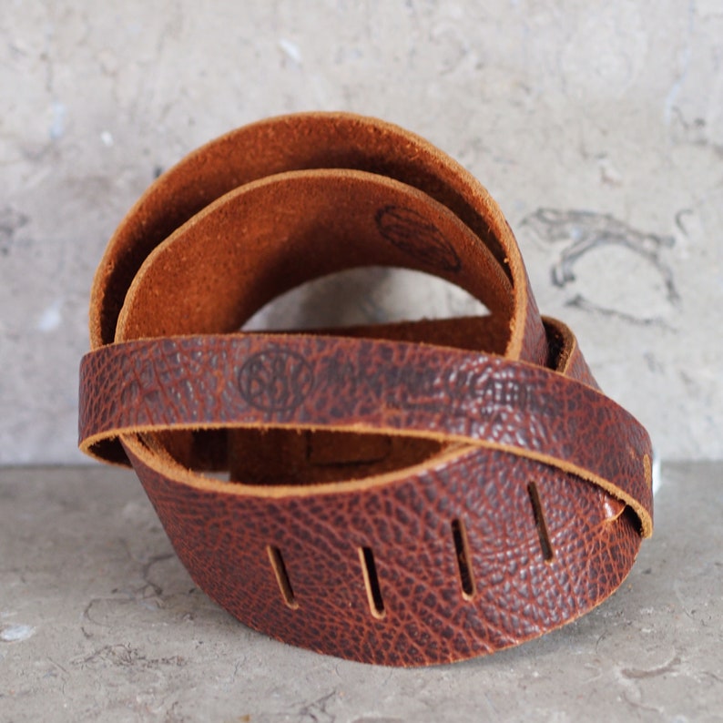 May include: A brown leather guitar strap with a textured surface and multiple holes for adjusting the length. The strap has a small logo embossed on it.