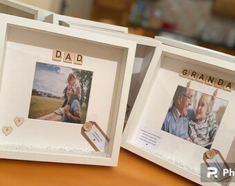 Può includere: Due scatole a ombre bianche con foto e tessere di lettere in legno che compongono le parole "DAD" e "GRANDA". Ogni scatola ha un piccolo cuore di legno e un'etichetta con un bottone.