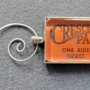 May include: A vintage-style keychain featuring a rectangular orange ticket with the text "CRESCENT PARK" and "ONE RIDE TICKET". The ticket is encased in a silver-colored frame and attached to a decorative silver swirl.