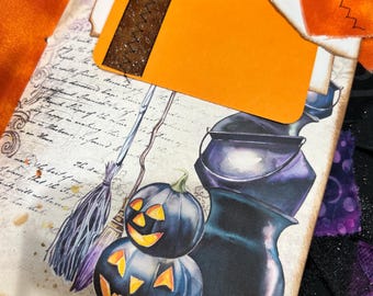 May include: A Halloween-themed journal with a decorative orange tag and a brown stitched ribbon. The journal cover features illustrations of jack-o'-lanterns, a witch's broom, and a cauldron. The background has vintage script and decorative fabric.