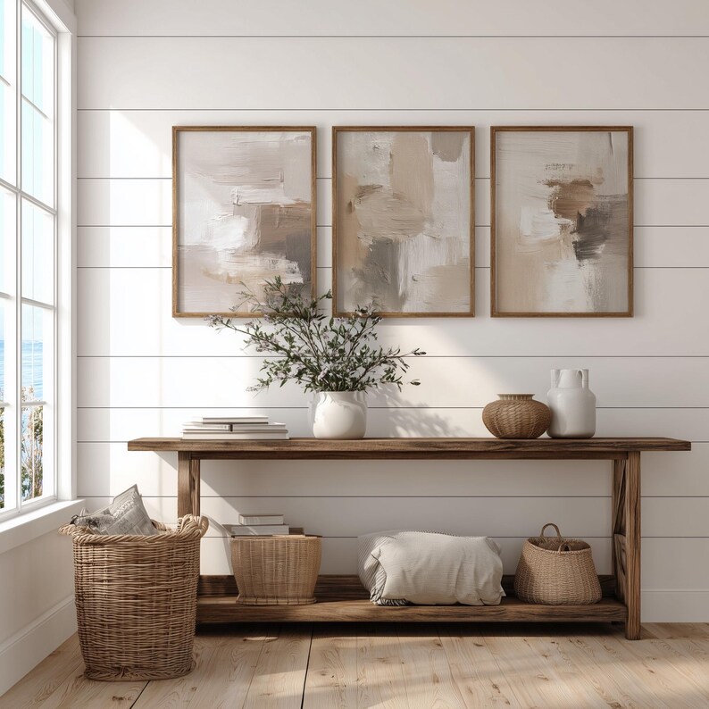 May include: Three framed abstract paintings in neutral tones hang above a wooden console table. The table holds a white vase with greenery, books, and decorative items. Woven baskets and a pillow are on the lower shelf.