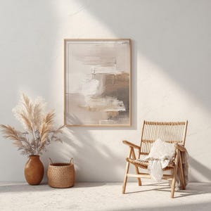 May include: Abstract art print in a wooden frame, featuring neutral tones of beige, white, and brown. The artwork hangs above a woven chair with a pillow and blanket. A vase with dried pampas grass and a woven basket complete the decor.