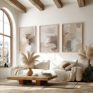 May include: Three framed abstract art prints in neutral tones hang above a white sofa. A wooden coffee table with a vase of pampas grass sits in front of the sofa. The room has a large window and wooden beams.