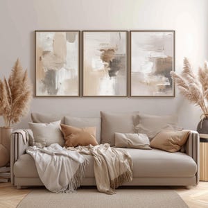 May include: Three framed abstract art pieces with neutral tones of beige, brown, and white hang above a beige sofa with throw pillows and blankets. Dried pampas grass in vases flanks the sofa, creating a warm, inviting living room scene.