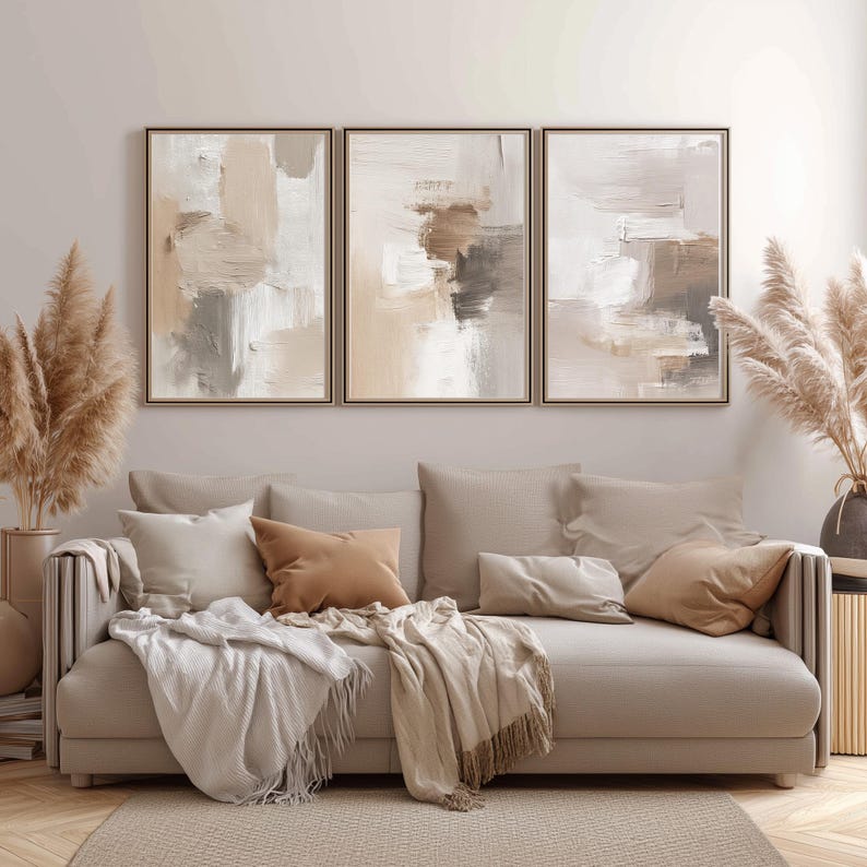 May include: Three framed abstract art pieces with neutral tones of beige, brown, and white hang above a beige sofa with throw pillows and blankets. Dried pampas grass in vases flanks the sofa, creating a warm, inviting living room scene.