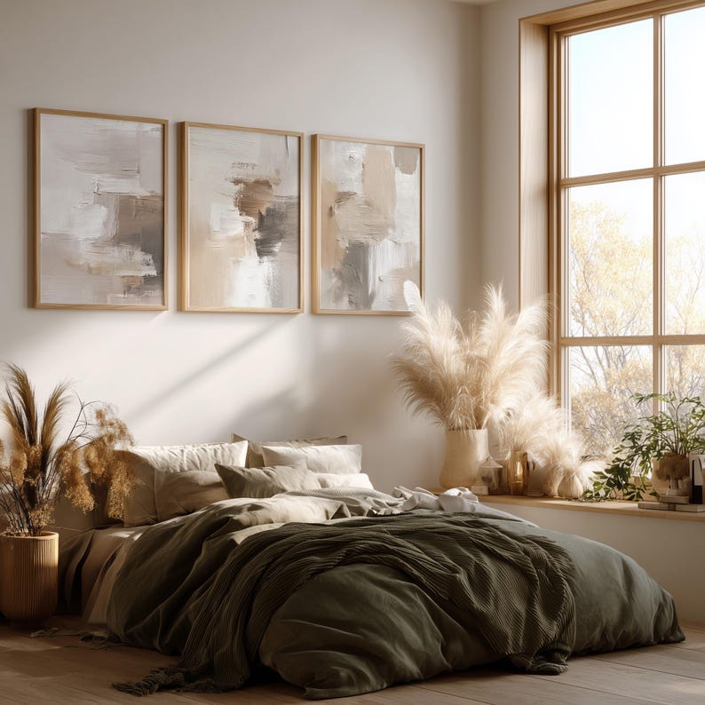 May include: A bedroom scene featuring a bed with olive green bedding and a neutral-toned comforter. Above the bed, three framed abstract paintings in neutral colors hang on the wall. The room is decorated with pampas grass and has a large window.