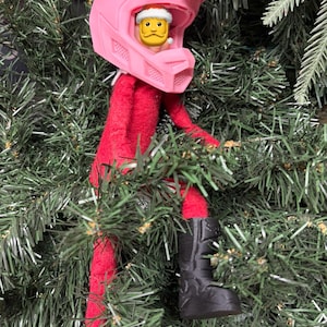 May include: A red elf figurine wearing a pink motocross helmet and black boots, nestled among green Christmas tree branches. The elf has a Lego Santa head. A fun and festive holiday decoration.