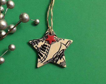 Pode incluir: Enfeite de Natal em forma de estrela com tema musical. O enfeite apresenta um fundo bege com notas e instrumentos musicais pretos. Uma pequena estrela vermelha é fixada no topo. Ele pende de um fio de barbante em um fundo verde.