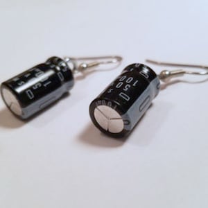 May include: A pair of earrings made from black cylindrical electronic components with silver hooks. The components have white text that reads "100 50".
