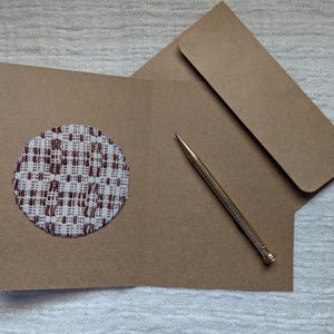 May include: A brown card with a circular fabric patch in a white and brown woven design. An envelope and a gold pen are also visible, suggesting a stationery set for correspondence.