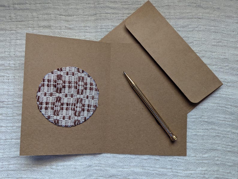 May include: A brown card with a circular fabric patch in a white and brown woven design. An envelope and a gold pen are also visible, suggesting a stationery set for correspondence.