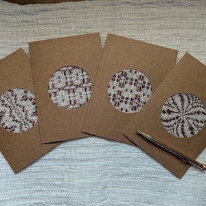 May include: Four brown greeting card envelopes with circular cutouts revealing patterned fabric inserts in white and burgundy. A gold pen rests on the surface. The cards are arranged on a white textured surface.