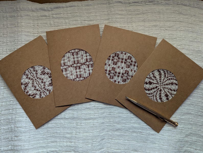 May include: Four brown card envelopes with circular cutouts displaying patterned fabric inserts in white and burgundy. A gold pen lies on the surface. The cards are arranged on a white textured surface.