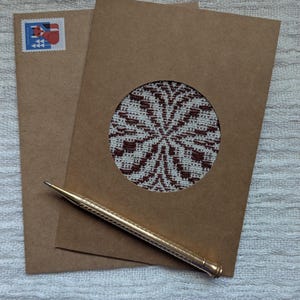 May include: Two brown paper cards, one with a circular cutout revealing a patterned design in white and brown. A gold pen rests on the cards, and a postage stamp is visible on the envelope. The cards and pen are on a white textured surface.