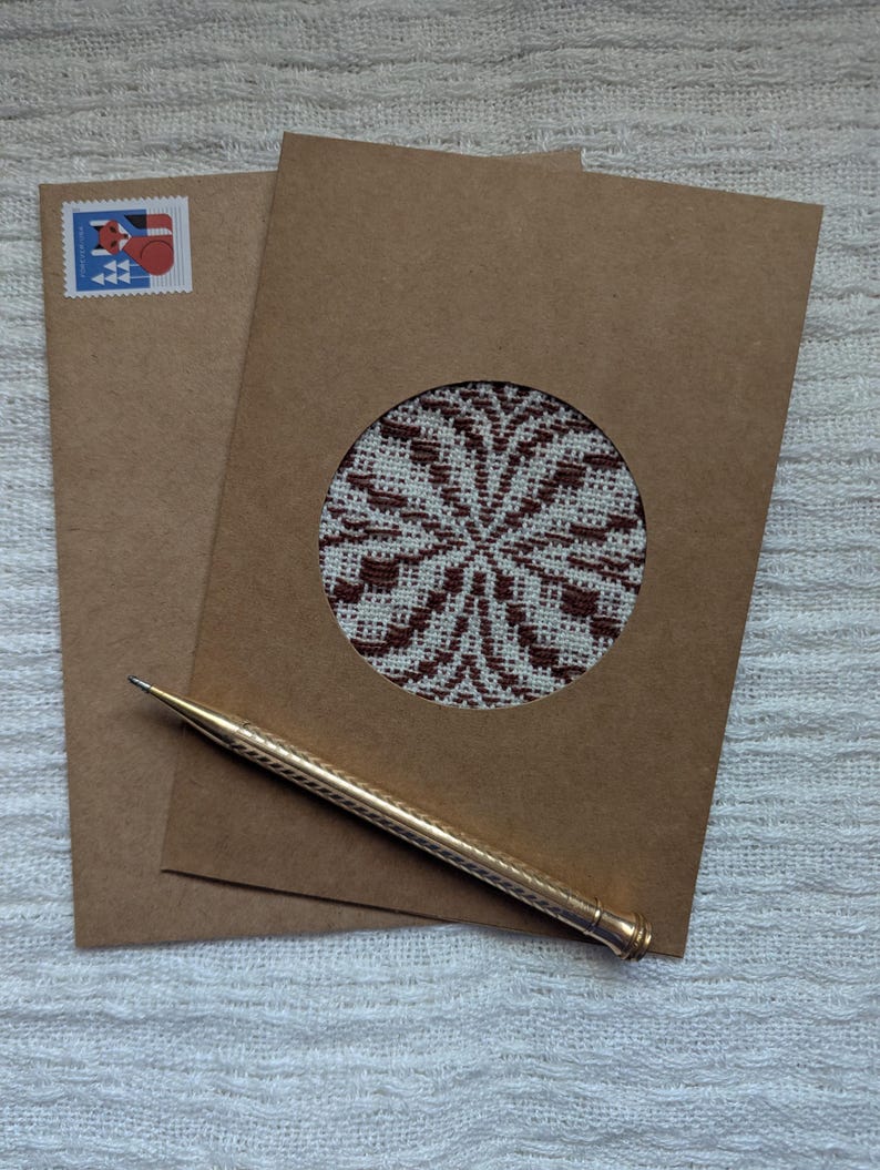 May include: Two brown paper cards, one with a circular cutout revealing a patterned design in white and brown. A gold pen rests on the cards, and a postage stamp is visible on the envelope. The cards and pen are on a white textured surface.