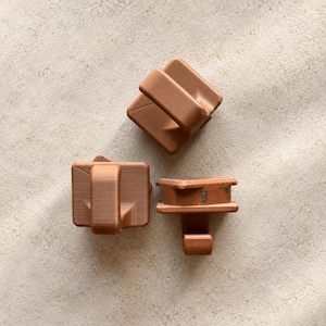 May include: Three copper-colored plastic hooks with a square base and a protruding J-shaped arm. Each hook features a smooth, matte finish with visible horizontal 3D printing layer lines on the surface of the material.