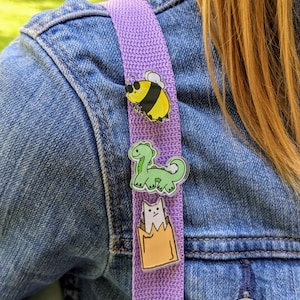 Op de afbeelding: Drie kleurrijke emaille pins bevestigd aan een paarse band. De pins tonen een geel-zwarte bij, een groene dinosaurus en een witte kat in een bruine zak. De band wordt over een spijkerjasje gedragen.