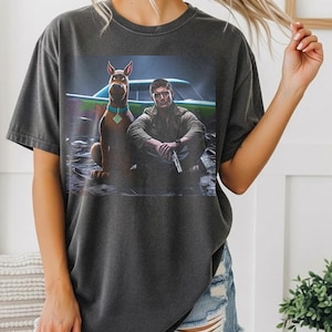 May include: Charcoal grey t-shirt with a graphic of a cartoon dog and a man with a car behind them. The dog wears a blue collar with a pendant. The man holds a gun. The text "PEPPER COMFORT COLORS 1717" is at the bottom.