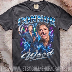 May include: A charcoal gray short-sleeve t-shirt featuring a graphic print of Connor Wood. The design includes the name Connor Wood in blue and white lettering, with multiple images of the person against a stage background with blue lightning.