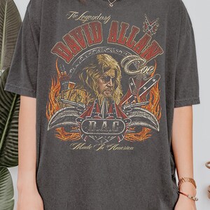 May include: Dark gray t-shirt featuring a graphic of David Allan Coe with text that reads "The Legendary David Allan Coe" and "Made in America." The design includes flames, guitars, and a D.A.C. logo.