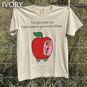 Könnte beinhalten: Ein elfenbeinfarbenes T-Shirt mit dem schwarzen Text 'You got a fast car. I got a plan to get us out of here.' und einer Grafik eines roten Apfels in Form eines Autos mit Rädern und Fenster, in dem ein rosa Wurm mit grünem Hut und roter Fliege sitzt.