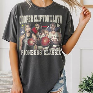 Puede incluir: Camiseta gris oscuro con un gráfico de tres jugadores de baloncesto sosteniendo balones. El texto "COOPER CLIFTON LLOYD" está encima de los jugadores y "PIONEERS CLASSIC" debajo. La camiseta es un Pepper Comfort Colors 1717.