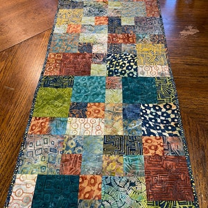 May include: A patchwork table runner with a variety of geometric and floral patterns in shades of blue, green, brown, and yellow.
