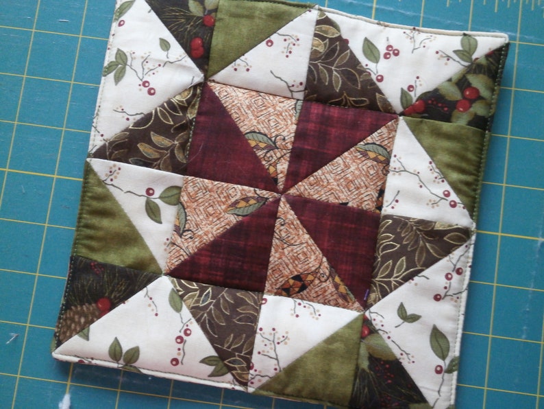 May include: A quilted fabric square with a starburst pattern in shades of brown, green, and white. The fabric features a leaf and berry design.