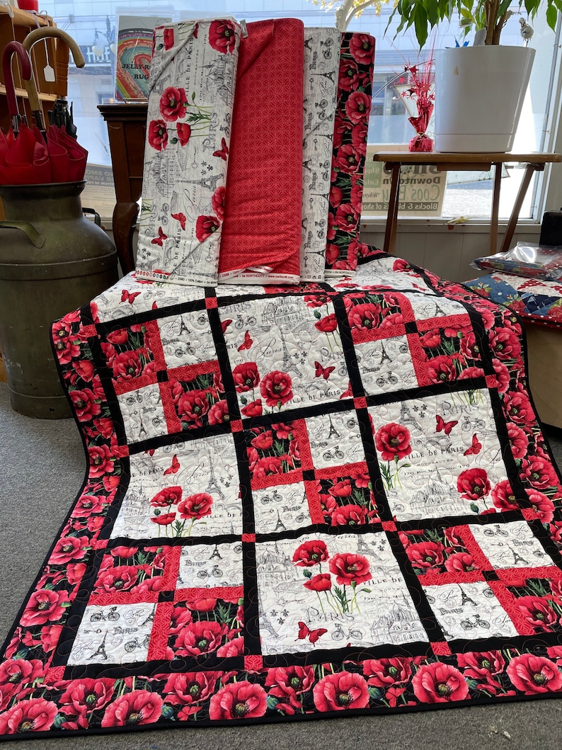 May include: A quilt with a black and white background featuring a repeating pattern of red poppies, bicycles, and the Eiffel Tower. The quilt has a black border with a red and white border inside the black border. The quilt is made of fabric scraps and is a patchwork design.