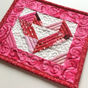May include: A pink and red quilted hot pad with a heart design in the center. The hot pad has a white background and a pink and red border with a swirl pattern.