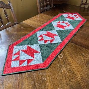 May include: A quilted table runner featuring a repeating pattern of red and white geometric shapes on a green background. The runner is bordered with a red trim and is displayed on a wooden table.