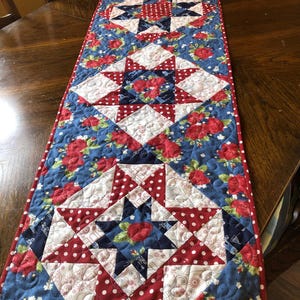 May include: A quilted table runner with a patriotic theme. The runner features a blue background with red roses and white polka dots. It has star and diamond patterns in red, white, and blue.