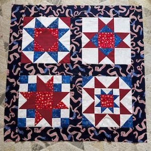 May include: A square quilt featuring four distinct star-patterned blocks in red, white, and blue. The quilt is bordered by a dark blue fabric with a repeating pattern of waving American flag ribbons and stars, showcasing a patriotic color scheme.