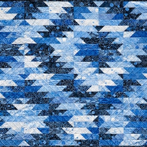 May include: A square quilt featuring a repeating geometric pattern in shades of navy, royal, and light blue. The fabric pieces display various intricate textures and small-scale prints, all joined by diagonal stitching lines across the surface.