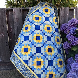 May include: A blue and yellow quilt with a star pattern. The quilt is folded and leaning against a wooden fence.