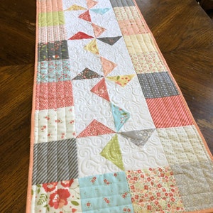 May include: A quilted table runner with a patchwork design featuring floral and geometric patterns in shades of pink, green, blue, and gray. The runner has a white background and a peach-colored border.