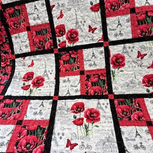 May include: A black and white quilt with a Parisian theme. The quilt features red poppies, Eiffel Towers, bicycles, and the word "Paris" repeated throughout. The quilt is made in a patchwork style with squares and rectangles.