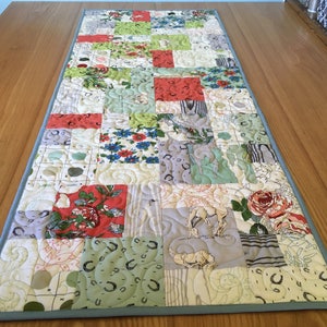 May include: A patchwork table runner featuring a mix of floral and geometric designs in shades of green, red, white and grey. The runner is quilted with a swirling pattern and has a light blue border.