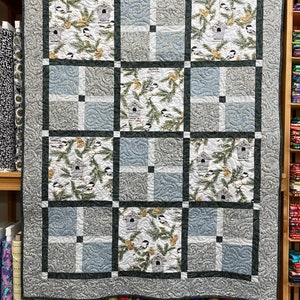 May include: A gray and white quilt with a black border. The quilt is made up of 12 squares, each with a different design. The designs feature birds, branches, and pine cones.