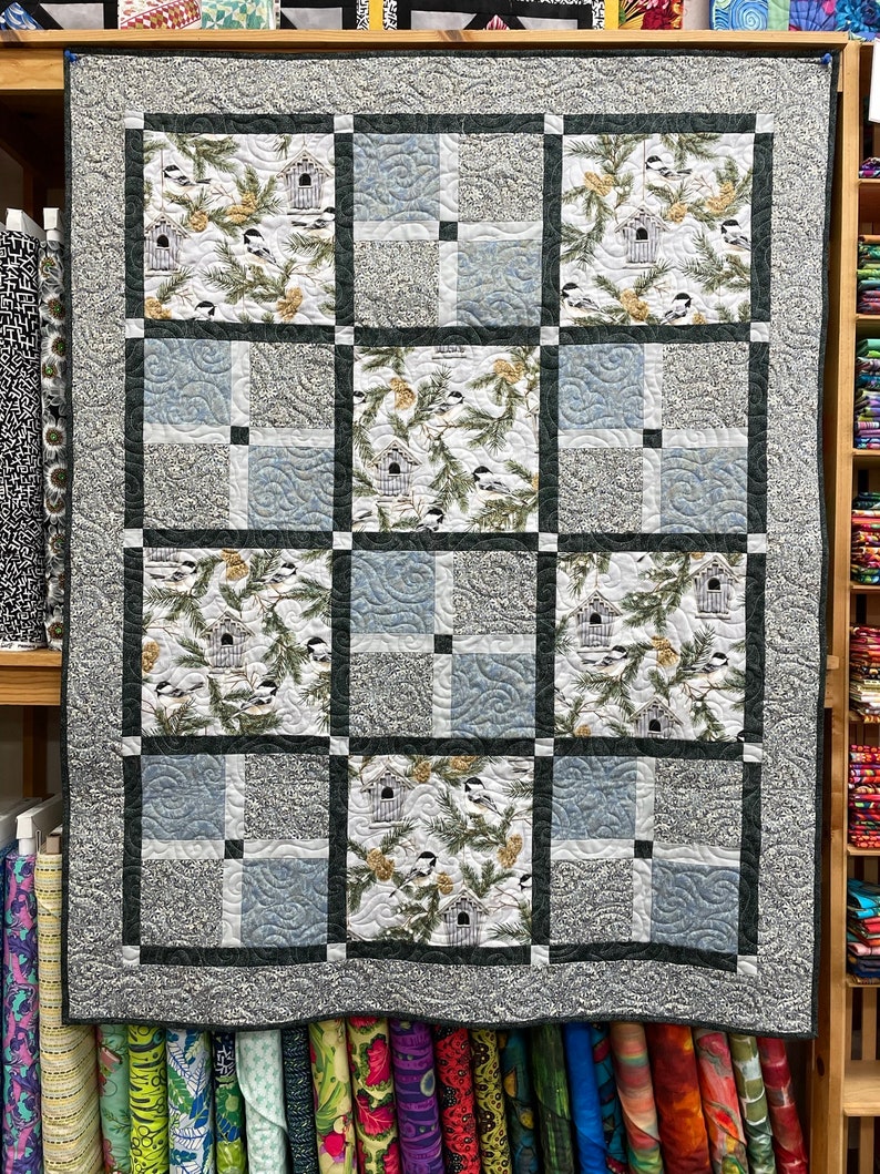 May include: A gray and white quilt with a black border. The quilt is made up of 12 squares, each with a different design. The designs feature birds, branches, and pine cones.