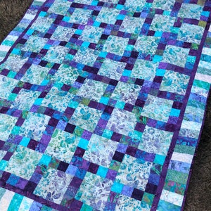 Könnte beinhalten: Eine Steppdecke mit Patchwork-Muster aus Quadraten in Lila, Blau und Weiß mit Blumenmuster. Die Steppdecke ist mit einem weißen und lila gestreiften Stoff eingefasst.