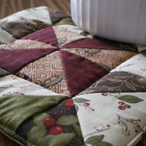 May include: A quilted pot holder with a patchwork design featuring various shades of green, brown, and red. The fabric has a floral pattern with small red berries.