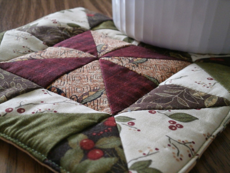 May include: A quilted pot holder with a patchwork design featuring various shades of green, brown, and red. The fabric has a floral pattern with small red berries.