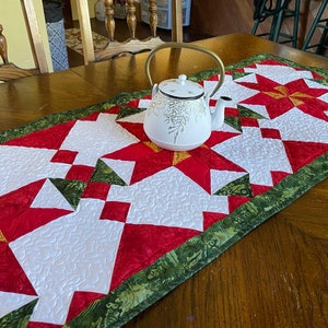May include: A quilted table runner with a red, white, and green star pattern. A white teapot with a gold handle sits on the runner. The runner has a green border with a leaf design. The table is made of wood.