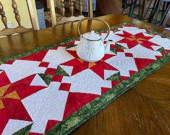 May include: A quilted table runner with a red, white, and green star pattern. A white teapot with a gold handle sits on the runner. The runner has a green border with a leaf design. The table is made of wood.