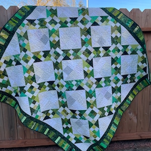 May include: A handmade quilt with a geometric pattern in shades of green and white. The quilt features square and diamond shapes, with a dark green border. The quilt is draped over a wooden fence.