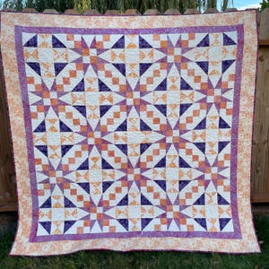 May include: A handmade quilt with a starburst pattern. The quilt features white, purple, and orange geometric shapes. The border is a patterned orange fabric. The quilt is displayed outdoors, draped over a wooden fence.