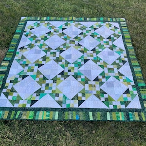 May include: A handmade quilt featuring a geometric pattern of white, green, and grey diamonds and squares. The quilt has a dark green border with a striped design. The quilt is lying on grass.