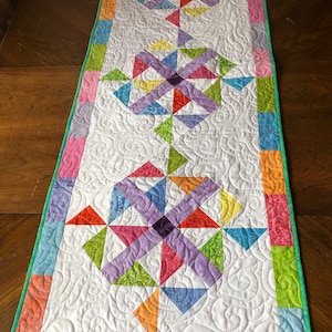 May include: A colorful table runner with a geometric pattern of triangles and squares in shades of purple, green, orange, blue, and yellow. The runner has a white background and a green border.