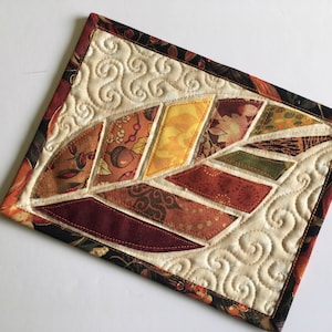 Può includere: Un sottobicchiere trapuntato con un design a foglia realizzato con ritagli di tessuto in tonalità di marrone, arancione, giallo e rosso. La foglia è circondata da uno sfondo bianco con un motivo a spirale.